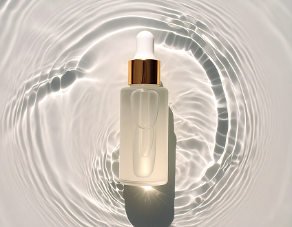 ゴールドのキャップ付きスポイトボトルが柔らかな光を受け、水の反射が広がるミニマルな化粧品イメージ